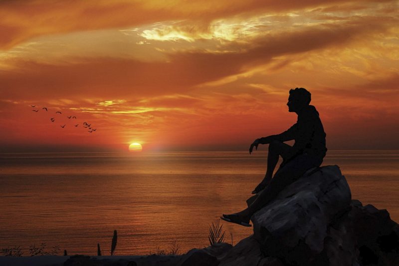 man sitting on boulder at the peach at sunset silhouette
